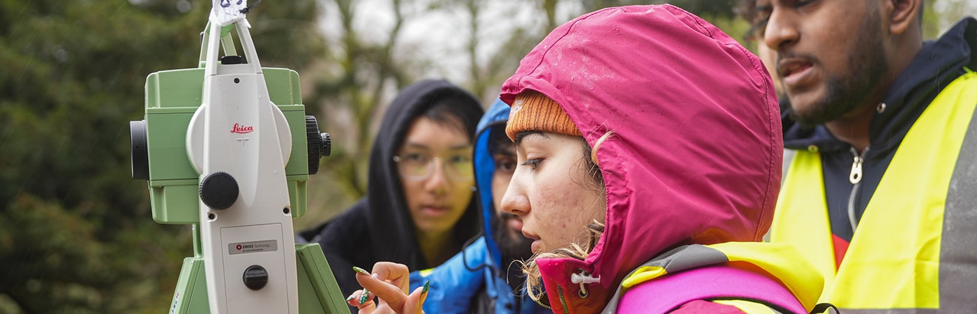 A woman student is using surveying equipment with other students in the background.